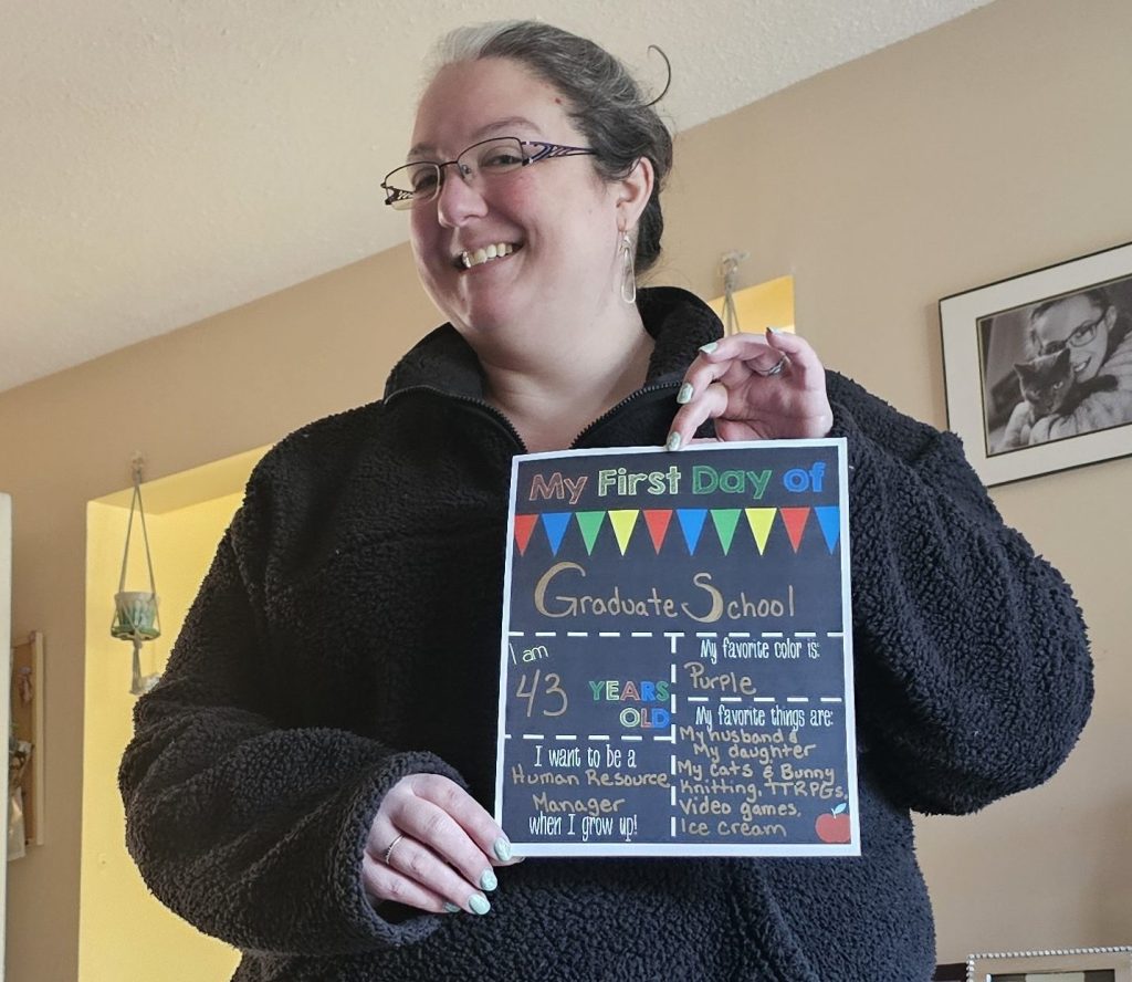 A woman with glasses and a black fleece jacket stands in a living room, smiling while holding a "My First Day of Graduate School" sign. The room has framed photos on a shelf, a backpack on the floor, and a partially open door in the background.