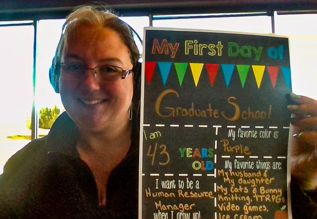 A woman with glasses and headphones smiles while holding a colorful sign that reads, "My First Day of Graduate School." The sign includes details such as her age (43), favorite color (purple), and favorite things. The background features large windows with natural light.