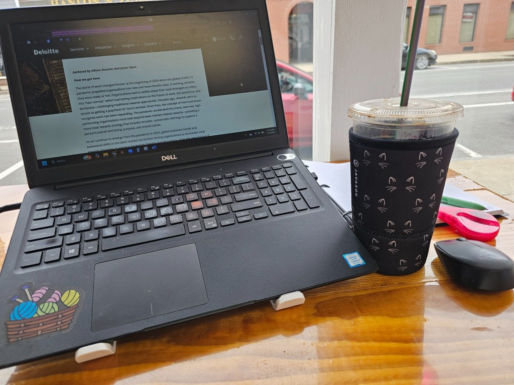 Laptop on a wooden table next to an iced coffee in a cat-themed sleeve, a computer mouse, pens, and highlighters, with a street view outside the window.