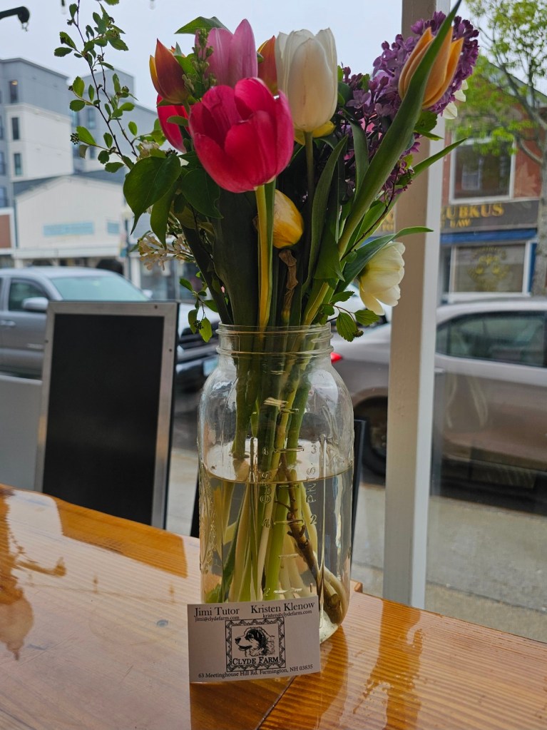 A glass mason jar filled with a colorful bouquet of spring flowers, including tulips and lilacs, sits on a wooden café table by a window. A business card for Clyde Farm, featuring the names Jimi Tutor and Kristen Klenow, is propped against the jar. Outside the window, parked cars and storefronts are visible on a cloudy day.