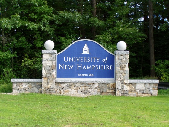 A large stone sign sits on a grassy area surrounded by tall green trees. The center of the sign features a blue panel with white text that reads “University of New Hampshire, Founded 1866.” Above the text is a simple white outline of a building with a steeple, representing a campus landmark.